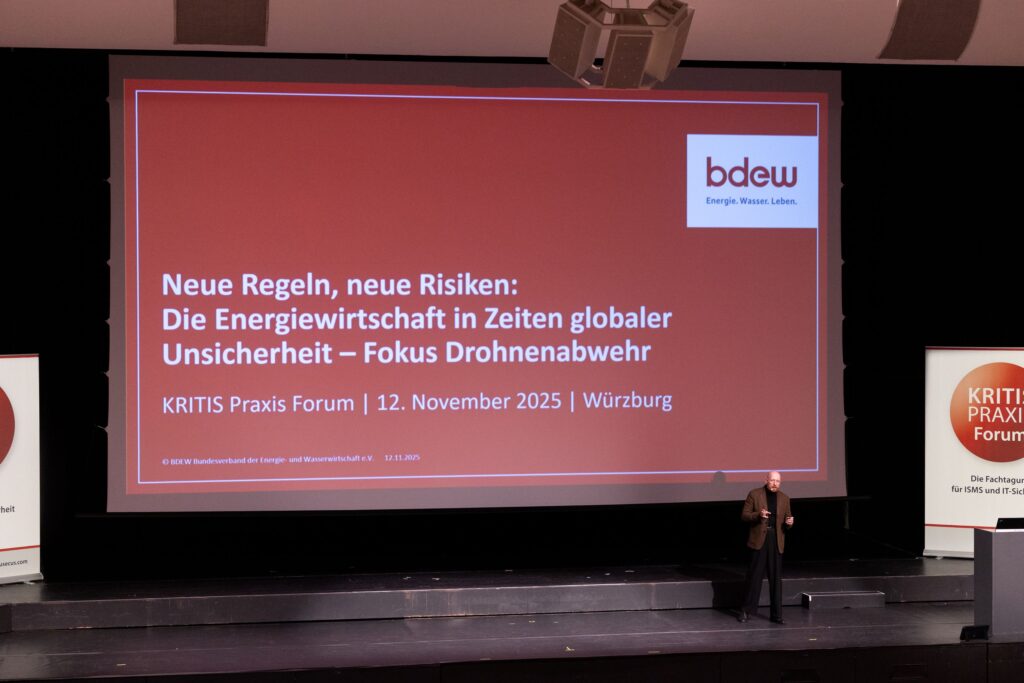 Mann spricht auf der Bühne vor einer Präsentation "Neue Regeln, neue Risiken: Die Energiewirtschaft in Zeiten globaler Unsicherheit – Fokus Drohnenabwehr" beim BDEW KRITIS Praxis Forum in Würzburg.