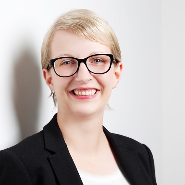 Nahes Porträt einer lächelnden blonden Frau mit Brille und schwarzem Blazer vor weißem Hintergrund.