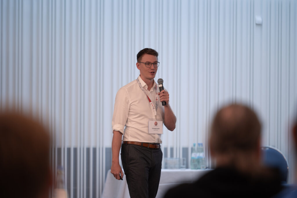 Christoph Knetti, ein junger Mann mit Brille und Hemd, spricht bei einer Konferenz ins Mikrofon vor Publikum.
