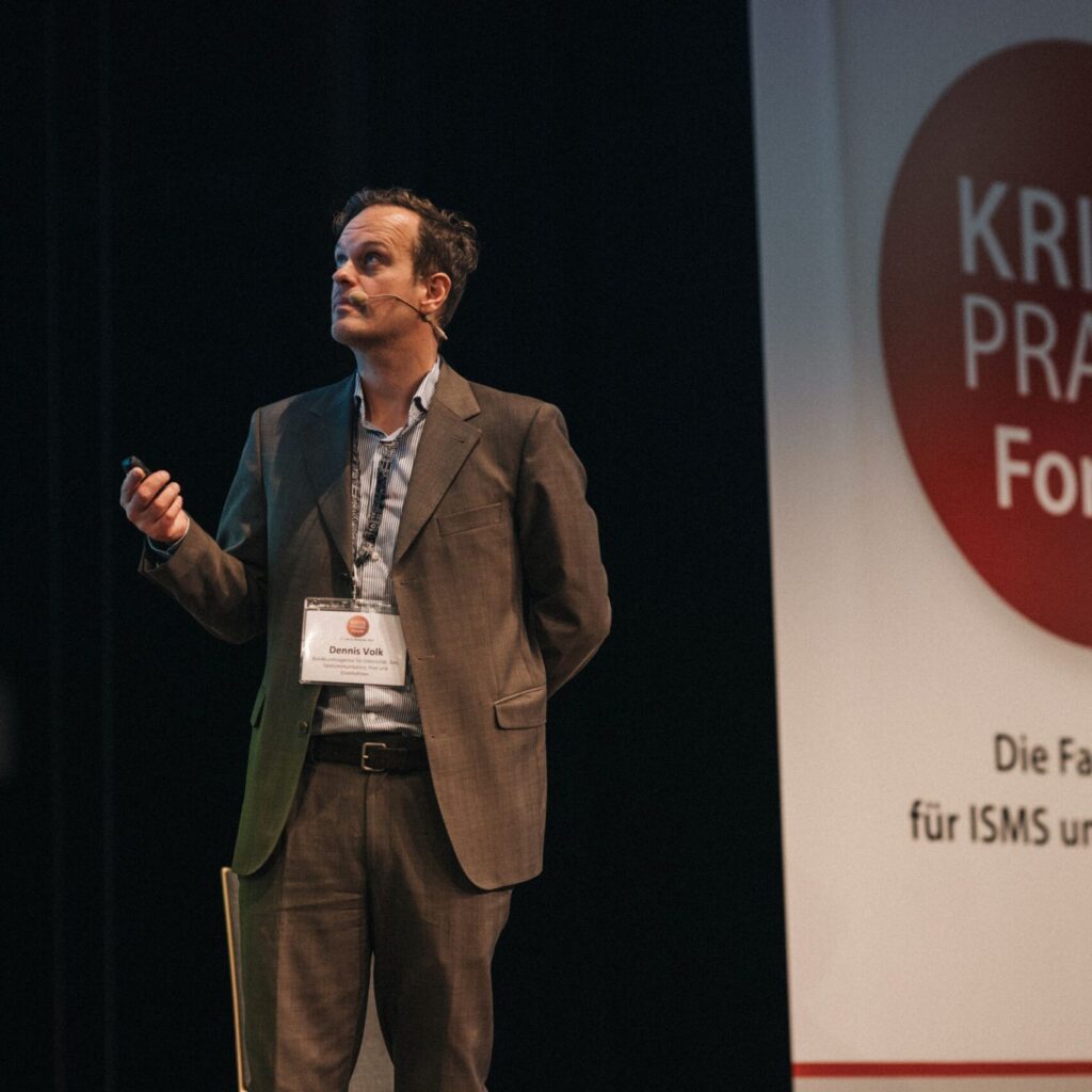 Dennis Volk, ein Mann im Anzug, präsentiert auf dem KRITIS PRAXIS Forum über ISMS und IT-Sicherheit.