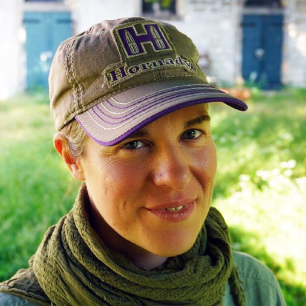 Eine lächelnde Frau mit Hornady Baseballkappe und olivgrünem Schal, im Freien, mit blauen Türen im Hintergrund.