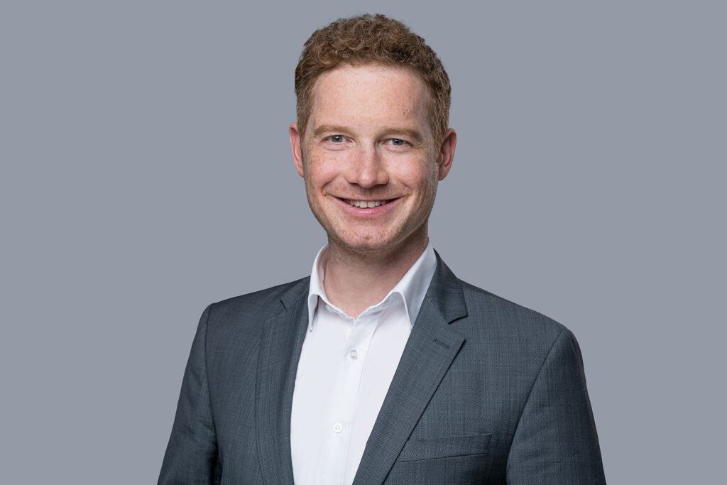 Freundlicher junger Geschäftsmann mit rotbraunem Haar und Anzug lächelt vor grauem Hintergrund.