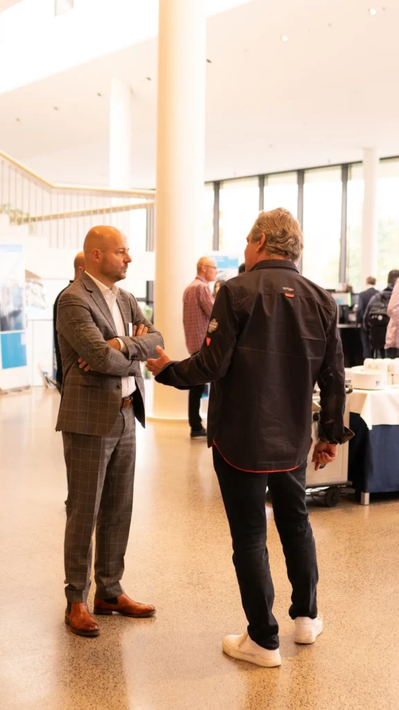 Zwei Männer unterhalten sich bei einer Konferenz, der eine trägt einen karierten Anzug, der andere eine schwarze Jacke. Im Hintergrund ist eine helle Lobby mit Fenstern zu sehen.