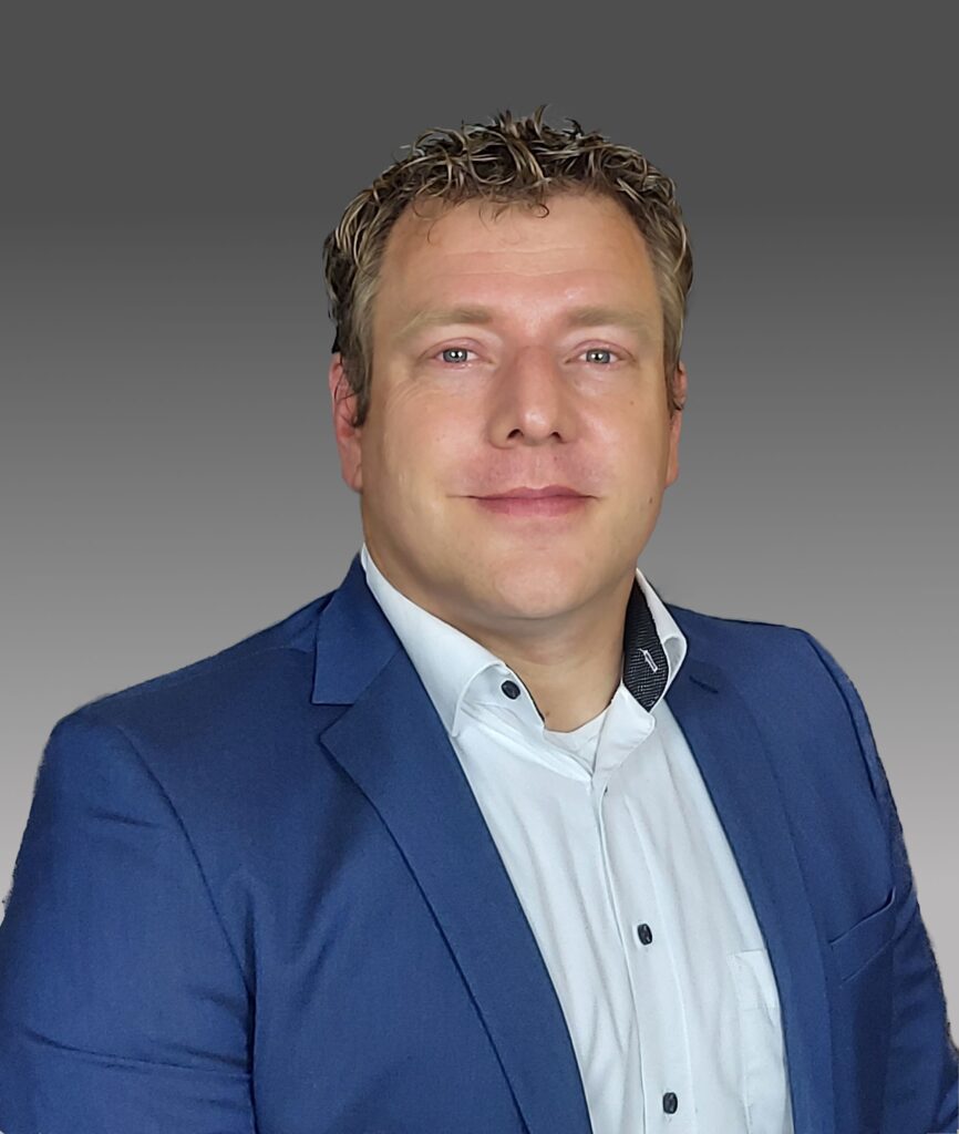 Porträt eines lächelnden Geschäftsmannes mittleren Alters mit blonden Locken, blauem Anzug und weißem Hemd vor grauem Hintergrund.