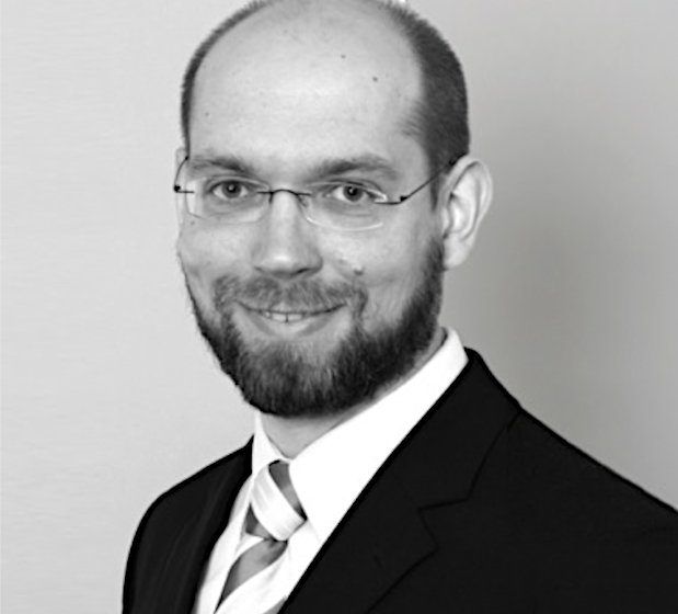 Schwarz-Weiss-Porträt eines lächelnden Geschäftsmannes mit Brille und Bart in Anzug und Krawatte.