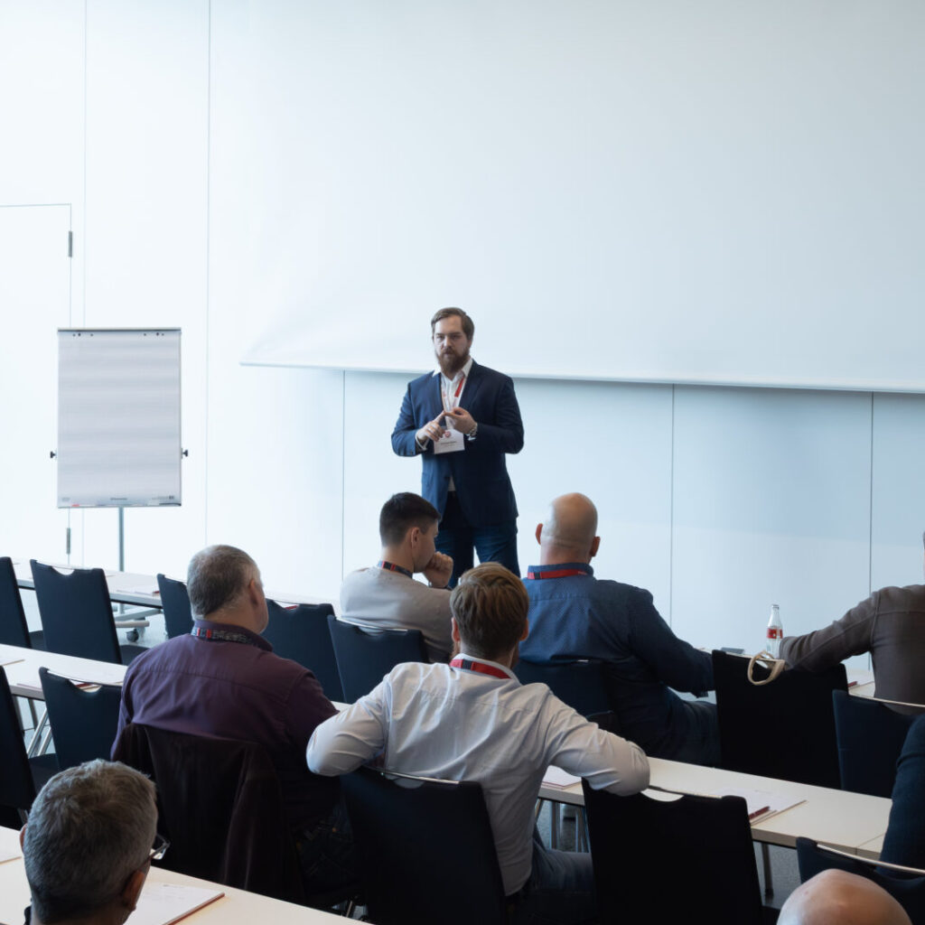 Ein Geschäftsmann hält eine Präsentation vor einem Publikum in einem modernen Konferenzraum mit großer Leinwand.