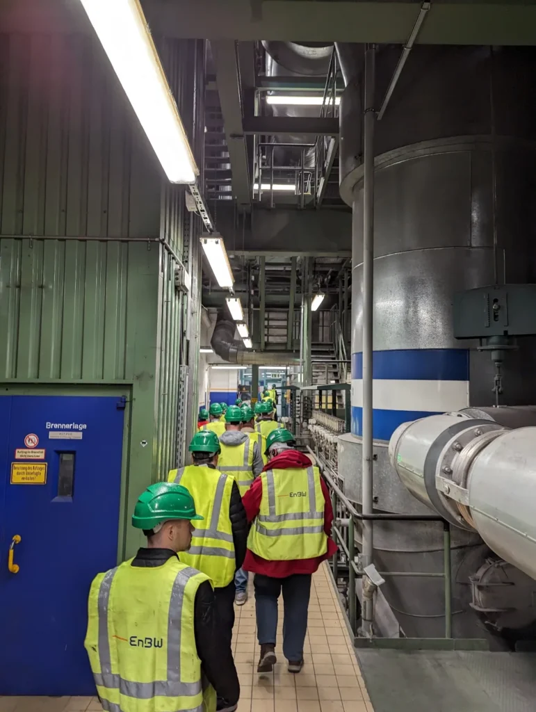 Gruppe von EnBW-Mitarbeitern in leuchtend gelben Sicherheitswesten und grünen Helmen bei einer Anlagenbesichtigung in einem Kraftwerk, umgeben von Industrieanlagen und Rohren.
