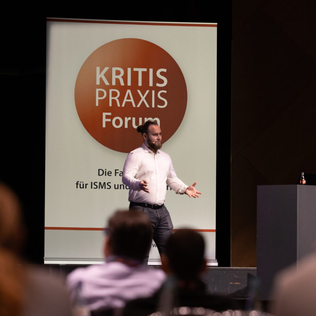 Ein Redner präsentiert auf der Bühne beim 'Kritis Praxis Forum' vor einem Publikum, mit Fokus auf Informationssicherheit.