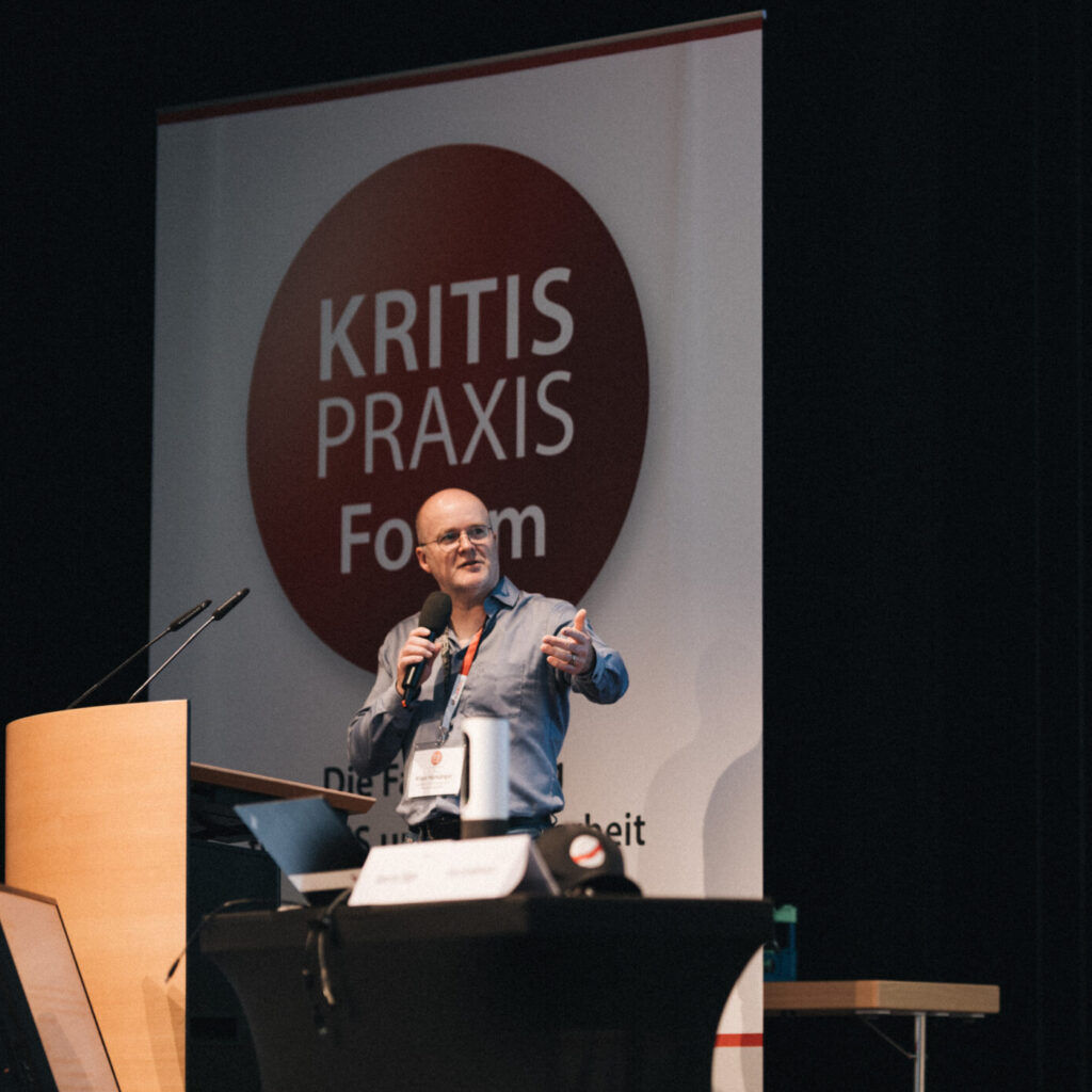 Zwei Männer auf einer Bühne bei einem Kritischen Infrastrukturen Praxis Forum; ein Vortragender spricht am Rednerpult.