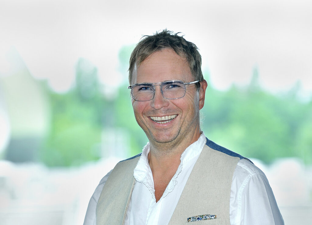 Ein lächelnder Mann mittleren Alters mit braunen Haaren, Brille und einer hellen Weste vor einem verschwommenen grünen Hintergrund.