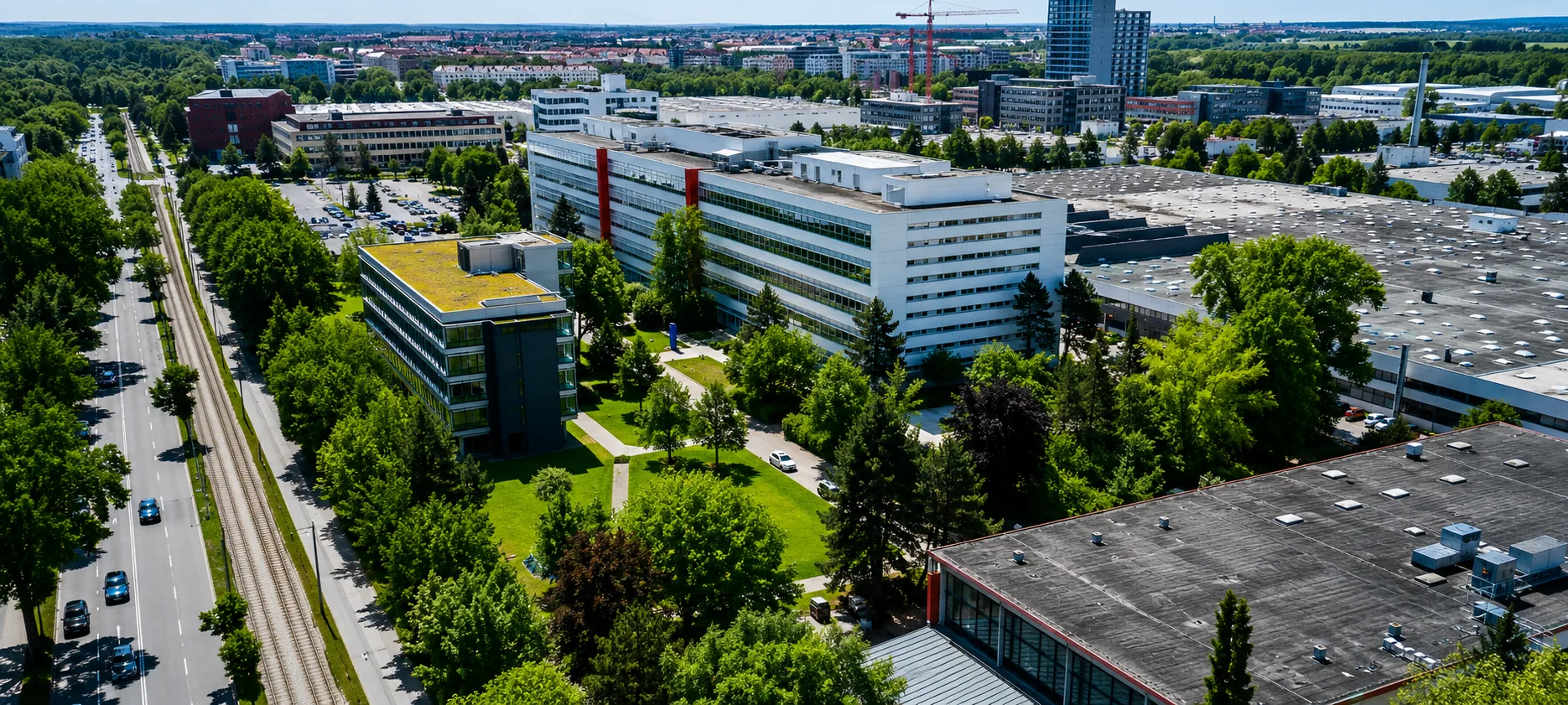 Luftaufnahme eines modernen Stadtgebiets mit Bürogebäuden, grünen Dächern, Straßen, Bahngleisen und üppigen Bäumen unter blauem Himmel.