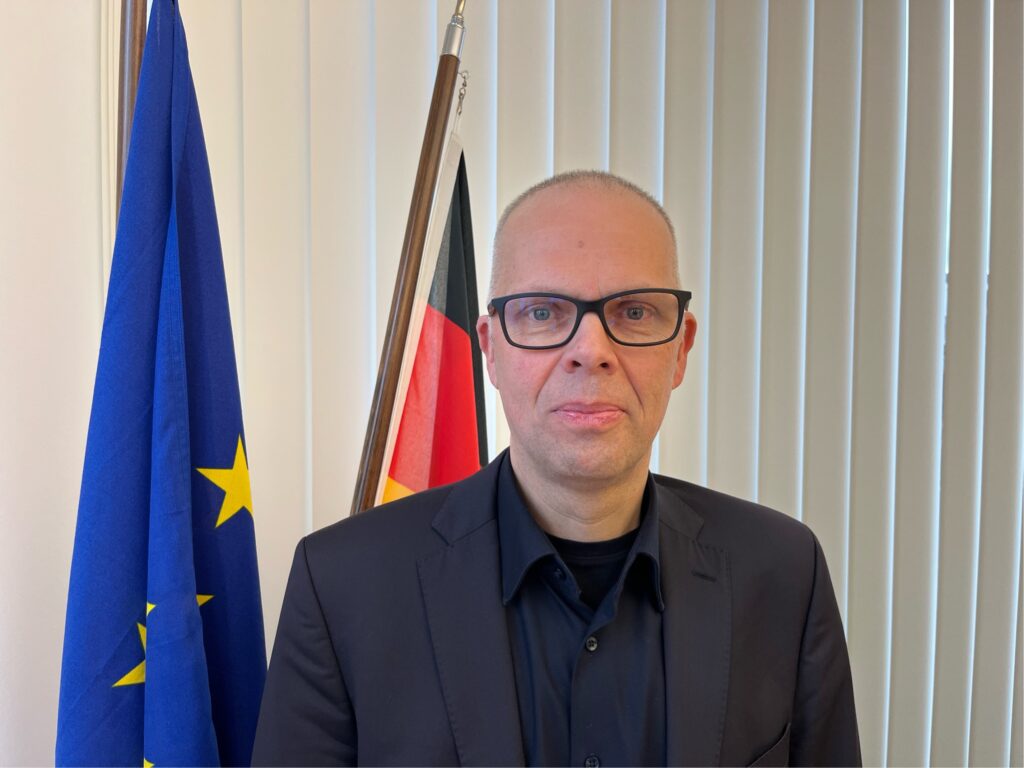 Ein Mann mit Brille und dunklem Anzug steht vor der EU- und der Deutschlandfahne.