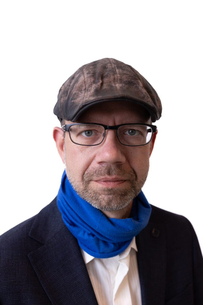 Porträt eines Mannes mittleren Alters mit brauner Schiebermütze, Brille, Bart und blauem Schal vor weißem Hintergrund.
