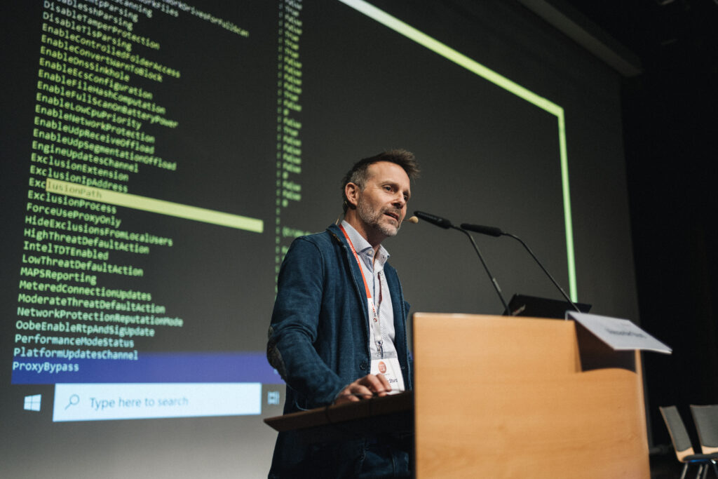 Ein Mann im Hemd und Sakko präsentiert auf einer Bühne vor einer großen Leinwand mit Code-Projektion.