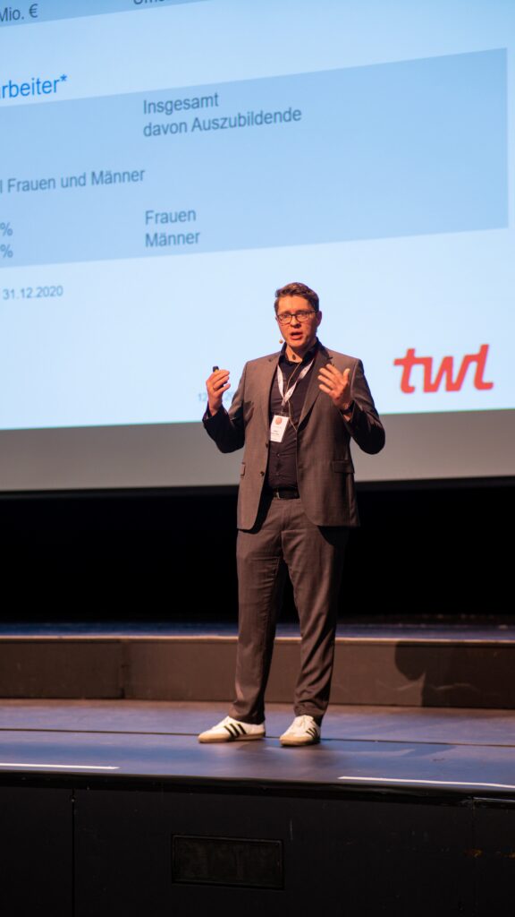 Ein Mann im Anzug präsentiert auf einer Bühne vor einer Projektionswand mit statistischen Daten und dem TWL-Logo.