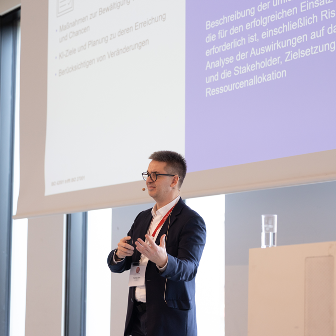 Ein Mann im Anzug hält einen Vortrag auf einer Konferenz mit einer Präsentation im Hintergrund, die KI-Risikobewertung und Ressourcenallokation zeigt.