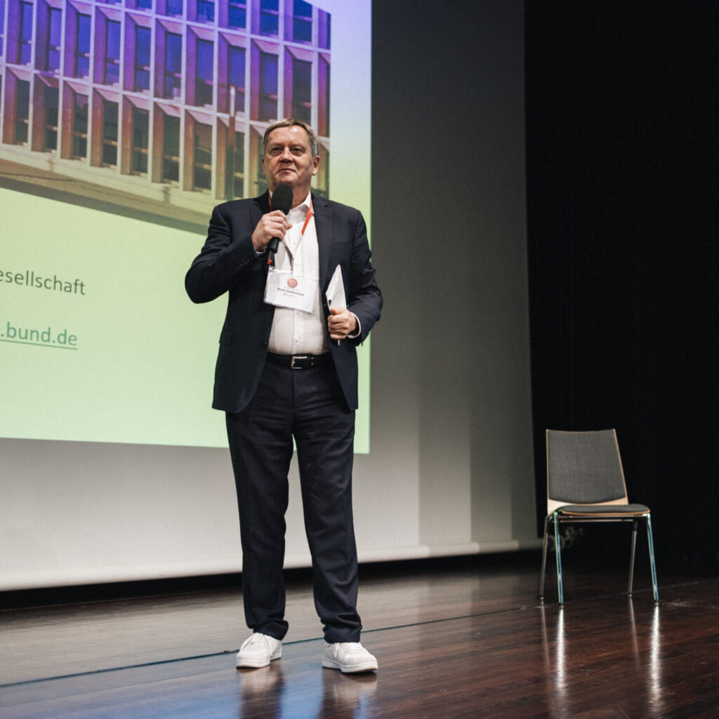 Ein Mann im Anzug präsentiert mit Mikrofon auf der Bühne des 'Kritis Praxis Forum', einer Fachtagung für ISMS und IT-Sicherheit.