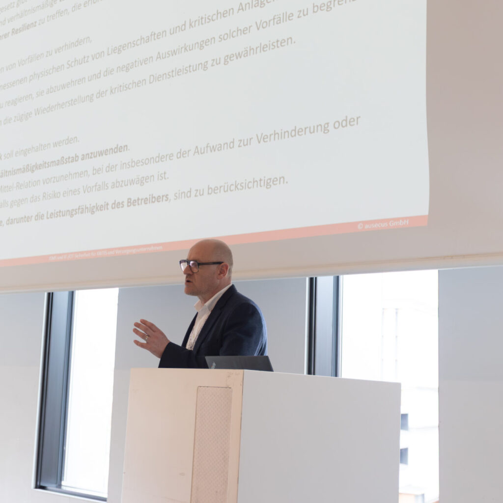 Ein Mann präsentiert vor einem Publikum in einem modernen Konferenzraum Details zum Resilienzplan und den Verpflichtungen für Betreiber kritischer Anlagen, mit Textprojektion im Hintergrund.