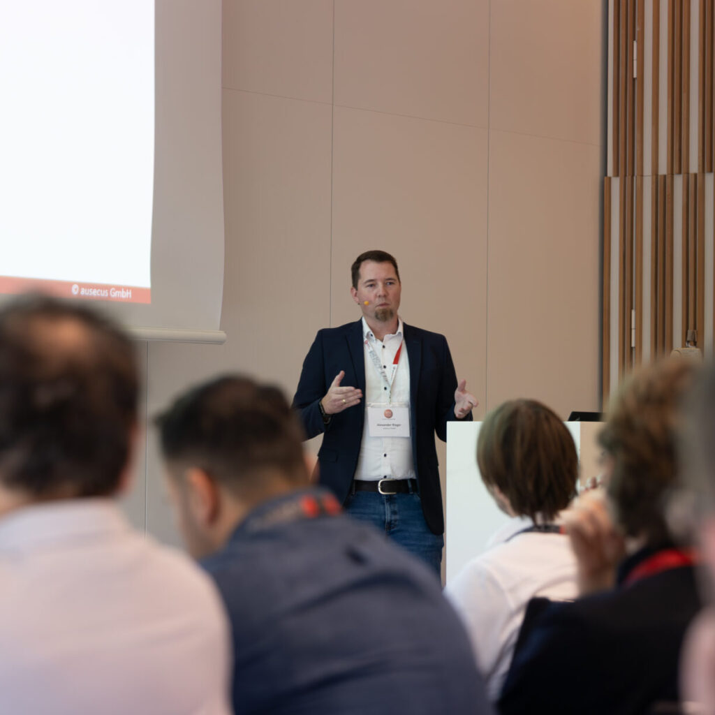 Ein Mann im Anzug präsentiert vor einem Publikum auf einer Konferenz, mit einer Projektionswand, die Text anzeigt.