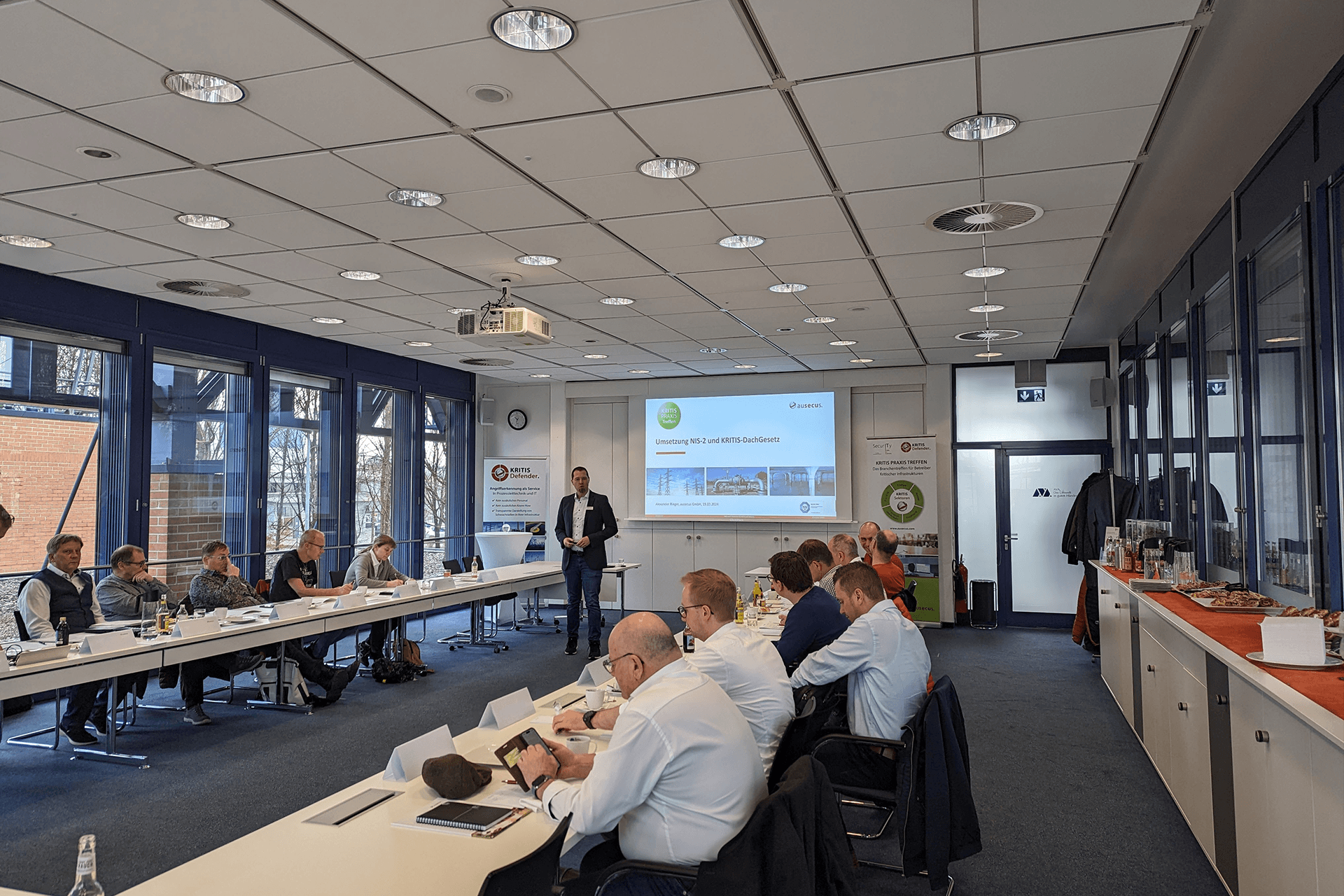 Ein Mann präsentiert vor einer Gruppe von Fachleuten die Umsetzung von NIS-2 und KRITIS-Dachgesetz in einem Konferenzraum.