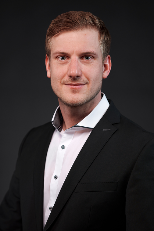 Porträt eines jungen, selbstbewussten Geschäftsmannes mit kurzem braunem Haar und Bartstoppeln, der lächelt und einen eleganten schwarzen Anzug mit weißem Hemd vor dunklem Hintergrund trägt.
