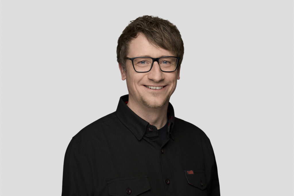 Porträt eines lächelnden Mannes mittleren Alters mit Brille und schwarzem Hemd vor grauem Hintergrund.