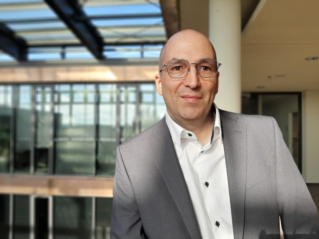 Porträt eines seriösen, glatzköpfigen Geschäftsmannes mit Brille und grauem Anzug vor verschwommenem Bürogebäudehintergrund.